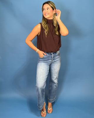 A woman modeling a sleeveless brown mock-neck top with light-wash blue jeans and metallic sandals