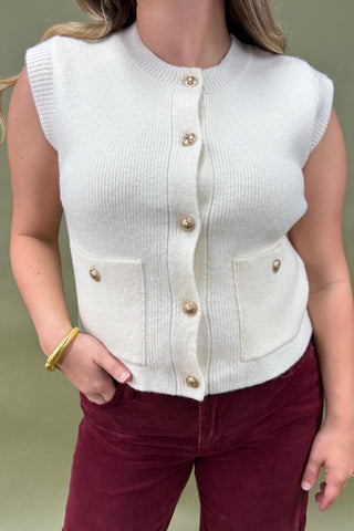 White sleeveless cardigan with gold buttons worn by a person on a plain background