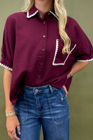 Person wearing a burgundy blouse with white trim and blue jeans against a green background