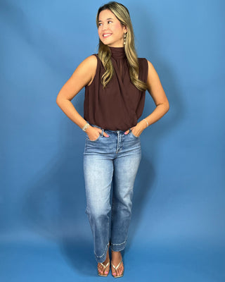 A woman modeling a sleeveless high-neck brown blouse and light-wash blue jean