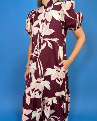 A woman wearing a short-sleeve tiered dress with a deep burgundy base and a large pale pink floral print, posed against a solid blue studio background.