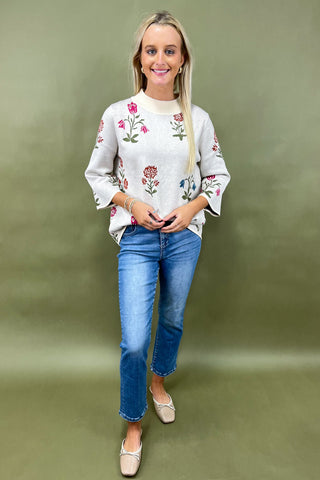 Woman wearing a white sweater with floral embroidery and blue jeans against a green background
