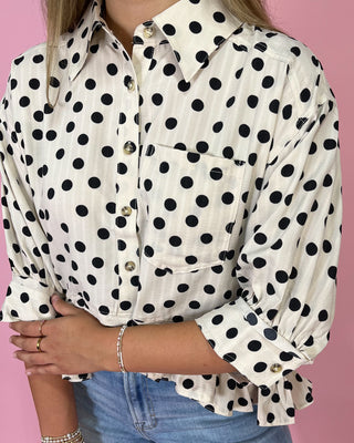 Person wearing a white shirt with black polka dots and blue jeans against a pink background
