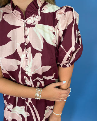 A close-up of a woman wearing a short-sleeve blouse with a burgundy and pale pink floral print, featuring a ruffled high collar and decorative buttons down the front. She is posed against a solid blue background.