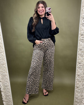 Woman taking a mirror selfie wearing a black top and leopard print pants against an olive green background.