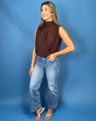 A woman modeling a sleeveless, high-neck, gathered brown top paired with light-wash blue jeans and flip-flop sandals