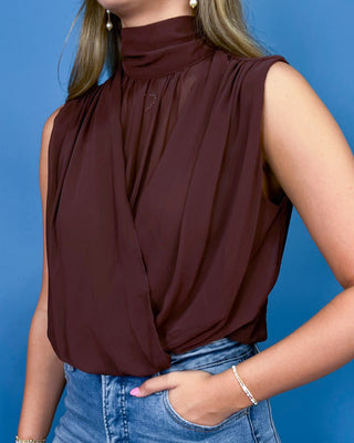 A woman wearing a sleeveless, high-neck, dark brown draped blouse tucked into blue jeans, set against a solid blue background.