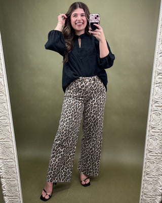 Woman taking a mirror selfie wearing a black top and leopard print pants on a green background.
