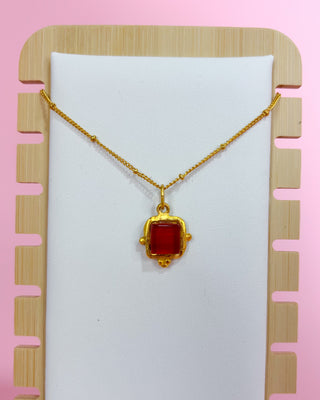 Gold necklace with a red pendant on a white card against a pink background