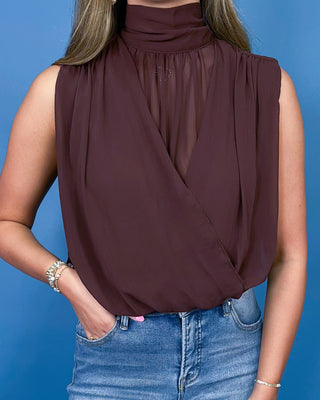 A woman wearing a sleeveless high-neck wrap-style blouse in a deep plum-brown color, paired with blue jeans, against a solid blue background.