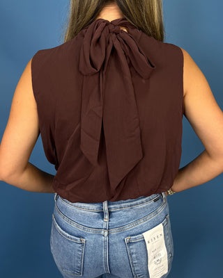 A woman viewed from the back wearing a sleeveless dark brown top with a large bow detail at the neckline, paired with light blue jean