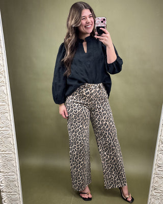 Woman taking a mirror selfie wearing a black top and leopard print pants on a green background