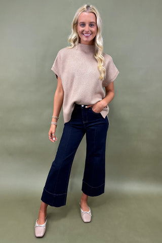 Woman wearing a beige top and dark blue jeans against a green background