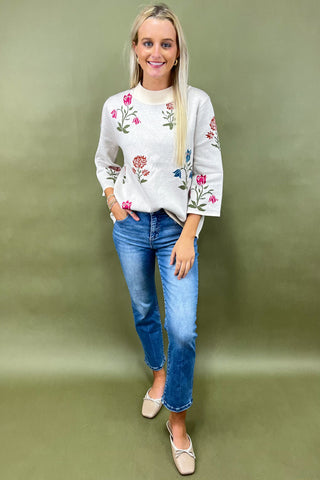 Woman wearing a white sweater with floral embroidery and blue jeans on a green background