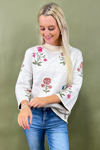 Woman wearing a white sweater with floral embroidery and blue jeans against a green background