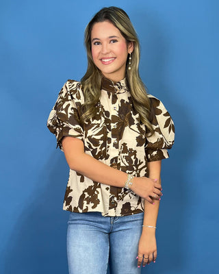 A young woman with blonde-highlighted hair smiling, wearing a brown and beige floral print blouse with puff sleeves and blue jeans
