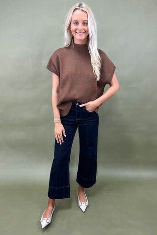 Woman wearing a brown top and dark jeans against a green background