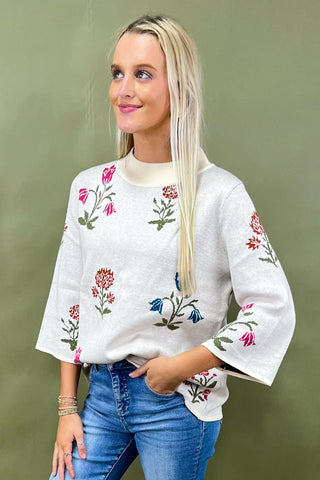 Woman wearing a white sweater with floral embroidery and blue jeans against a green background