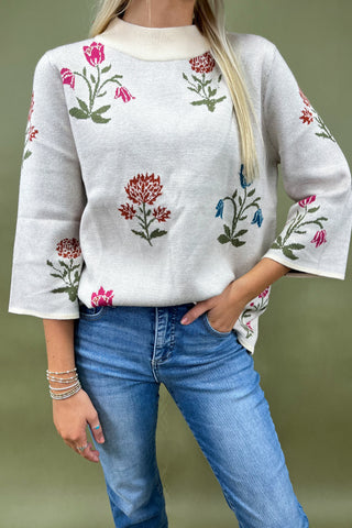 Woman wearing a white sweater with floral embroidery and blue jeans against a green background