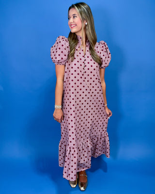 A woman modeling a pink midi dress with dark red polka dots, featuring puffed short sleeves and a ruffled hem, set against a solid blue studio background.