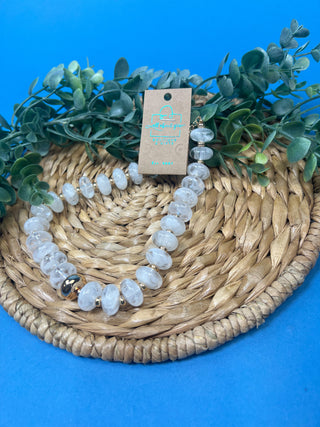 A beaded necklace made of clear, polished oval beads with a small gold-toned accent bead, displayed on a woven straw mat with green leaves against a bright blue background. A kraft paper tag is attached to the necklace.