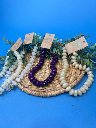 Three beaded necklaces displayed on a woven tray with greenery against a bright blue background. One necklace is deep purple, and two are translucent off-white.
