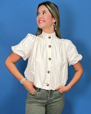 A woman wearing a cream-colored short-sleeve blouse with a high mock neck and decorative gold buttons, paired with olive green jeans