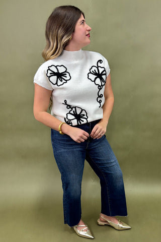 Woman wearing a white top with black floral patterns and blue jeans against a green background