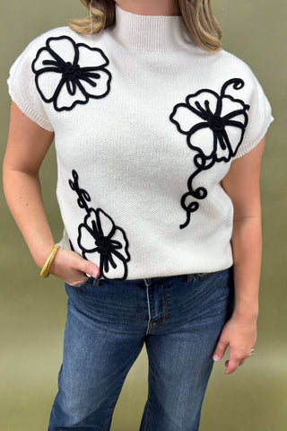 White sweater with black floral patterns worn by a person on a beige background