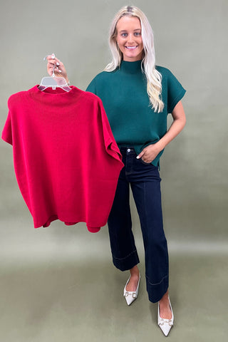 Woman wearing a green top and dark jeans on a plain background holding a red top