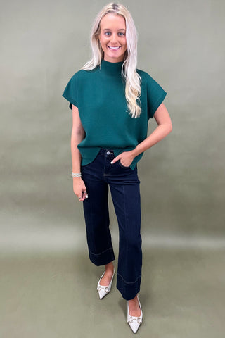 Woman wearing a green top and dark jeans on a plain background