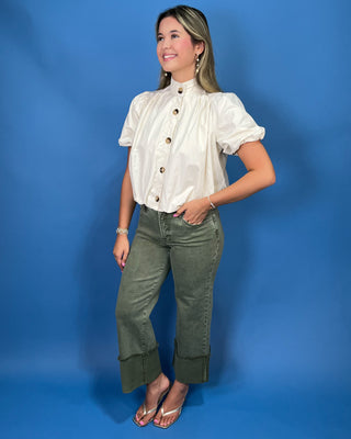 A woman modeling a cream-colored short-sleeve button-up blouse with puff sleeves and olive green cropped jean