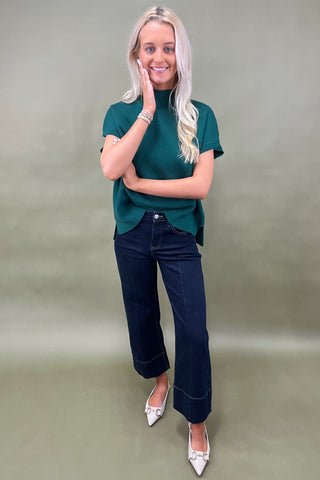 Woman wearing a green top and dark jeans on a plain background