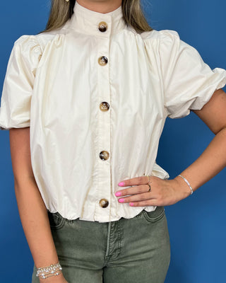 A woman wearing a cream-colored short-sleeve blouse with a high mock neck and decorative gold buttons, paired with olive green jean
