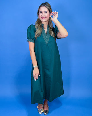 A woman modeling a dark teal green midi dress with puff sleeves and a V-neckline, standing against a solid blue studio backdrop.