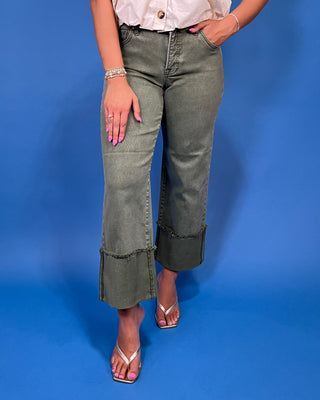 A model wearing a pair of olive green wide-leg cropped jeans with rolled cuffs, paired with a white blouse