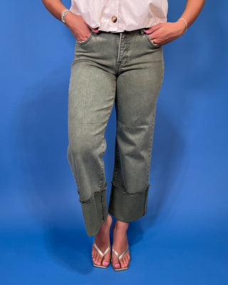 A person wearing faded sage green cropped jeans with rolled cuffs and frayed hem