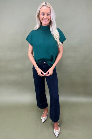 Woman wearing a green top and dark jeans on a plain background