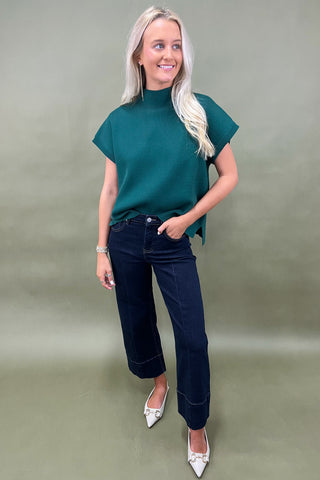 Woman wearing a green top and dark jeans on a plain background