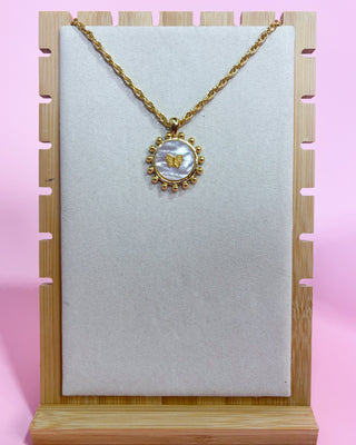 Gold necklace with a circular pendant on a white card, against a pink background