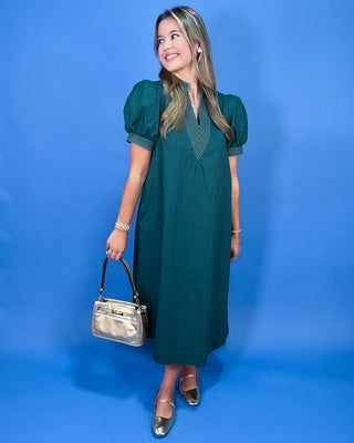 A woman modeling a dark green midi dress with puff sleeves and a striped V-neckline, holding a metallic silver handbag against a solid blue background.