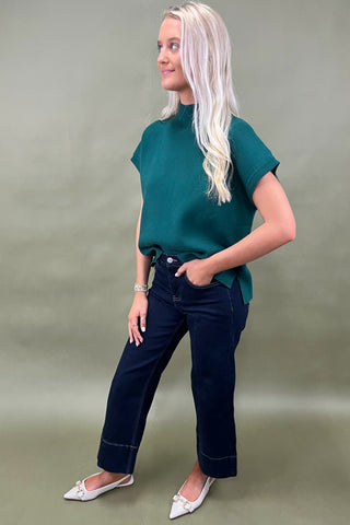 Woman wearing a green top and dark jeans on a plain background