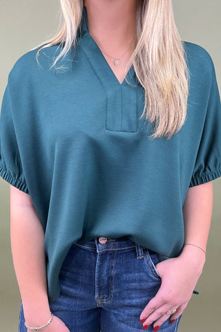 Person wearing a teal blouse with a pocket and blue jeans against a neutral background