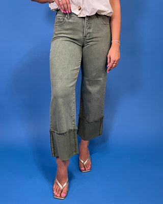 A model wearing a pair of olive green wide-leg cropped jeans with rolled cuffs, paired with a white blouse