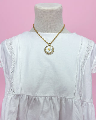 White dress with lace details and a gold necklace on a mannequin against a pink background