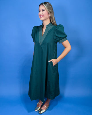 A woman modeling a dark green midi dress with puff sleeves, a V-neckline, and striped trim details, standing against a solid blue studio backdrop.