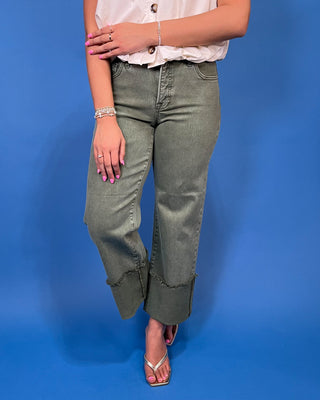 A model wearing a pair of olive green wide-leg cropped jeans with rolled cuffs, paired with a white blouse