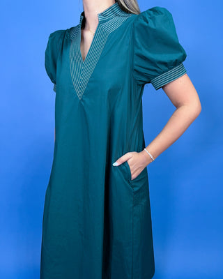 A woman wearing a teal green short-sleeve dress with puff sleeves, a striped V-neckline, and side pockets, posed against a solid blue background.
