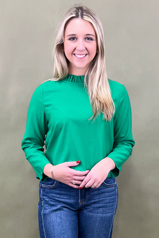 Woman wearing a green long-sleeve top and blue jeans against a beige background