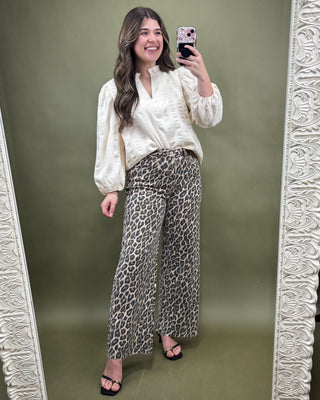 Woman taking a mirror selfie wearing a white blouse and leopard print pants against a green background.